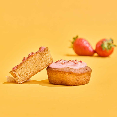 Two sugar-free No Guilt Bakes Strawberry Swirl | Keto Cake Bite | Limited Edition | 10 Cakes rest on a yellow background; one is cut in half displaying its interior, while two whole strawberries are blurred in the background.