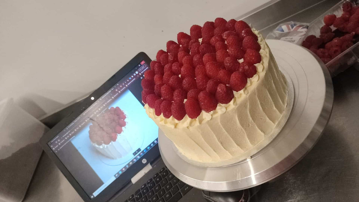 Custom Made Low Sugar Cakes from No Guilt Bakes: A delectable masterpiece, adorned with freshly picked raspberries, is elegantly showcased on a table only inches away from a sleek laptop. Our skilled bakers specialize in fulfilling your custom cake creation.