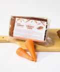 A package of low-carb, keto-friendly Carrot Keto Cake Loaf and fresh carrots on a cutting board by No Guilt Bakes.