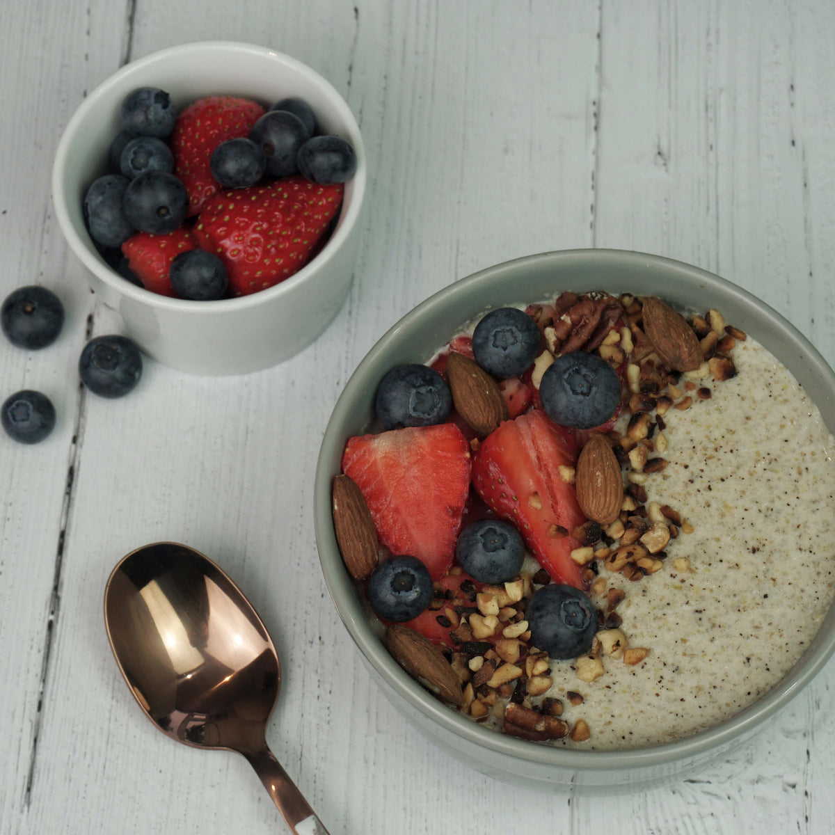 A bowl of chia pudding topped with strawberries, blueberries, almonds, and granola, accompanied by a spoon and a small bowl of mixed berries on a wooden surface. Ideal for a low-carb diet. Made with No Guilt Bakes&#39; Regular Bread Bundle.
